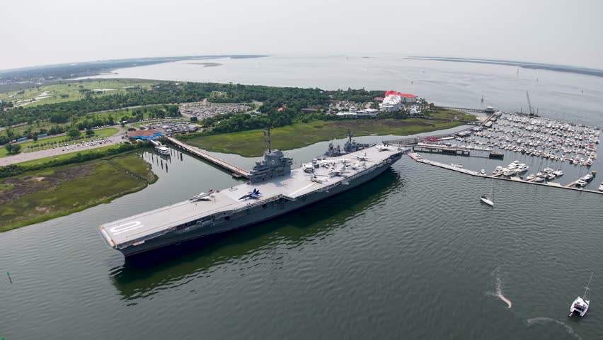 A sweeping drone shot of the USS Yorktown aircraft carrier moored at Patriots Point in Charleston Harbor showing the surrounding water and coastline.