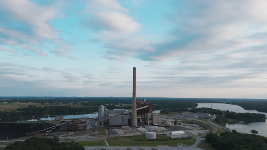 Drone establishing pullback from smokestack of power plant next to wide lake inlet bordered by forested hills, peaceful sky
