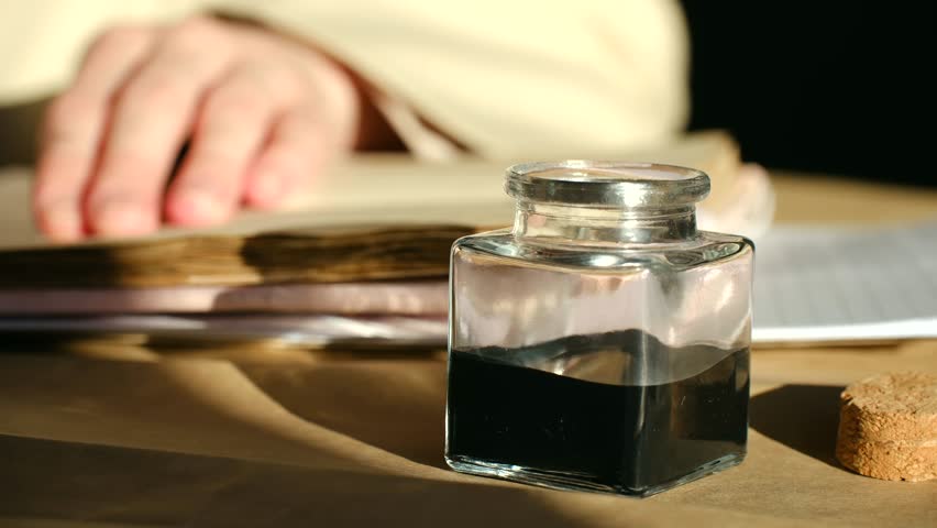Close up writer hand dipping classic nib pen into glass inkwell filled with black ink, preparing to write on old paper in sunlit room, evoking a historical, scholarly mood. 