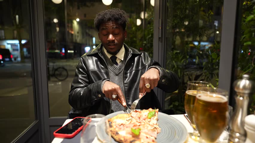 Confident african american man sitting alone at a restaurant table by the window, cutting a slice of pizza for dinner while enjoying a beer and looking at the camera in the evening