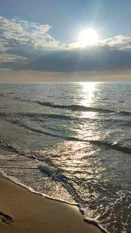 Seascape on sunny summer day. Sea waves rolling on sand of sandy beach on seashore. Waves of calm sea, sand, blue sky with clouds and horizon line. Sun shining from behind rainy clouds. Vertical