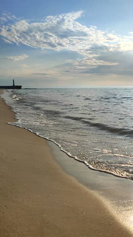 Seascape on sunny summer day. Sea waves rolling on sand of sandy beach on seashore. Waves of windless calm sea, blue sky with clouds and horizon line. Nature backdrop. Natural background. Vertical