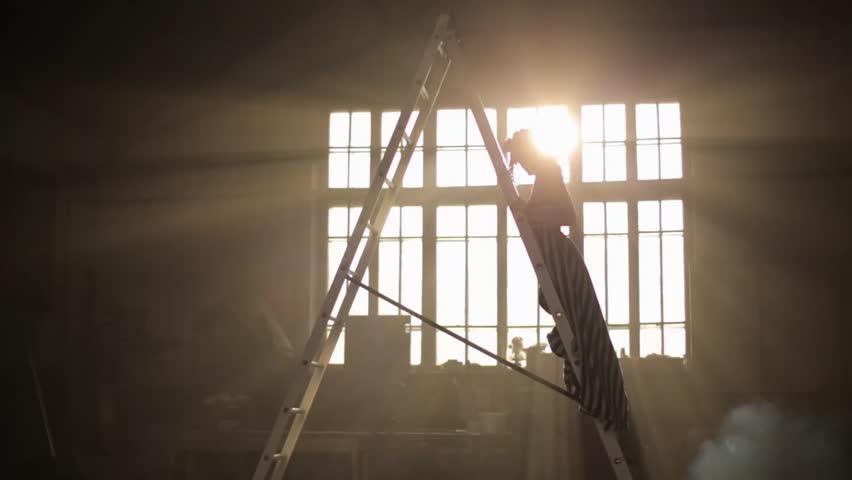 A powerful silhouette of a woman standing on an industrial ladder in a dark, smoky warehouse. Dramatic sun rays burst through a large window, symbolizing effort, challenge, or aspiration