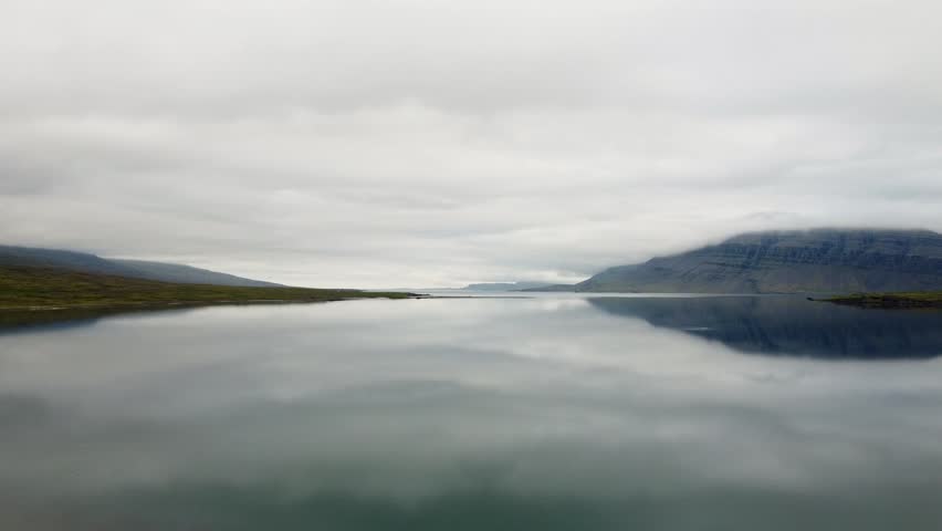 Serene aerial footage of a mist-shrouded lake with reflective calm water and adjacent hillside cabins. Captured near Berufjörður Fjord, this tranquil Icelandic vista evokes a sense of peace.