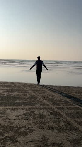 Silhouette of a young man jumping on the wet sand at the beach during sunset, with the ocean waves gently rolling in. He is in mid-air, arms spread wide, capturing a moment of freedom and joy against 