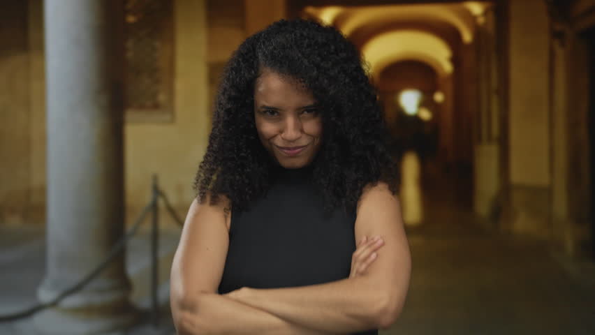 Young hispanic woman sticks tongue out in old town street archway while crossing arms; playful rebellion.