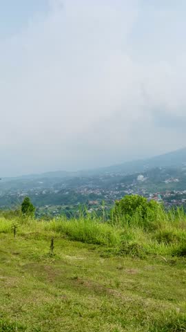 Panorama of rural landscape showing lush green meadows in the foreground. The forest area leads to a valley filled with small buildings. Mountains rise in the distance. The sky is mostly cloudy