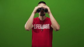 Man holds binoculars to eyes while wearing lifeguard shirt and whistle against a green studio backdrop, arms visible and focused; vigilance duty. - Powered by Shutterstock - Get 15% off with code: PIKWIZARD15