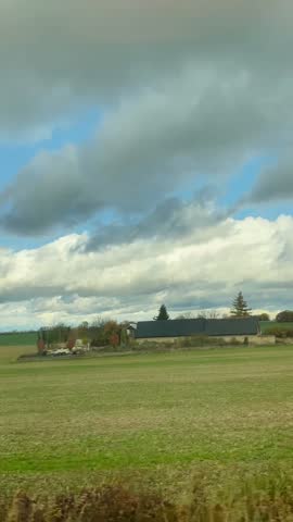 Vertical Dramatic Clouds Over Open Farmland Fields, Blurred Trees Sweep Past During Transit, Distant Farmhouse And Horizon Line Visible, Traveling Passenger Capturing Stormy Scene on mobile device