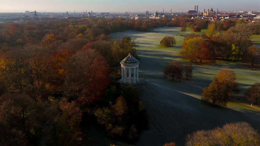 Drone footage of Englischer Garten in Munich at dawn, capturing Monopteros and colorful autumn trees. In distance are the towers of Frauenkirche, Theatinerkirche and the Bavarian State Chancellery