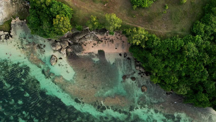 Overhead view of Bali beach displays transparent reef water, sandy shore and tall coastal cliffs as shallow waves move in steady lines across the tropical shoreline under clear sky.