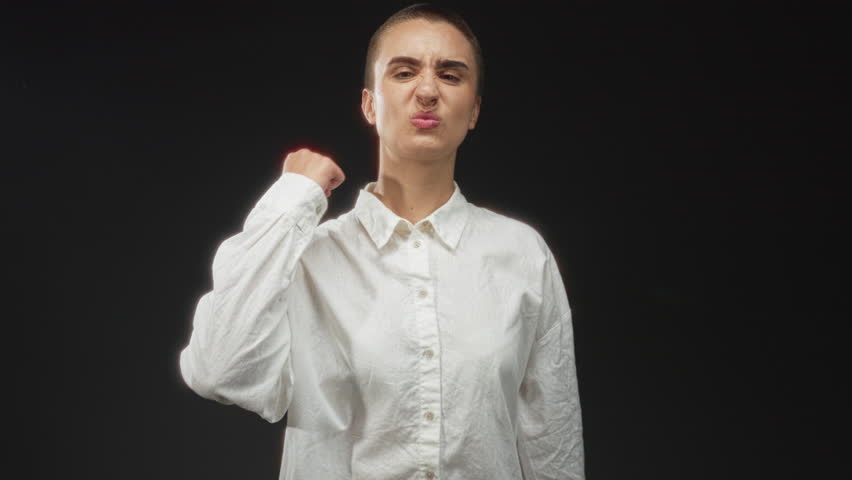 Young woman with clenched fist raised toward camera, grimacing with bared teeth, wearing white shirt in studio; defiance determination.