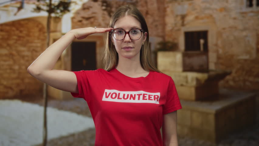 Woman saluting on a cobblestone street by historic stone facade wearing a volunteer shirt and eyeglasses; duty respect.