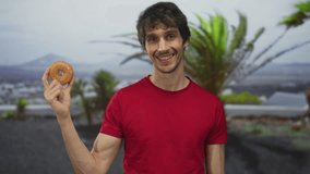 Man wearing red shirt holding glazed doughnut with fingers and smiling cheerfully on street; joy celebration. - Powered by Shutterstock - Get 15% off with code: PIKWIZARD15