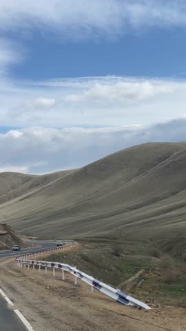 Scenic winding mountain road with cars under a cloudy sky, showcasing vast, rolling hills and distant horizons. A journey through a tranquil, expansive landscape.