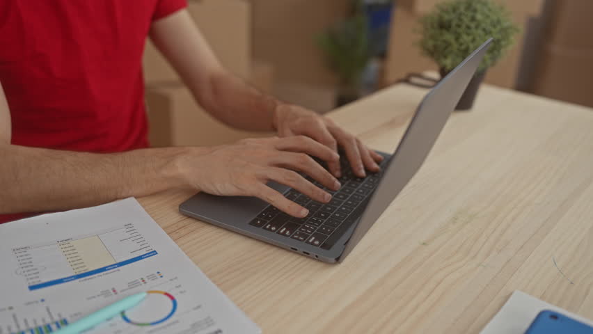 Young man in red shirt typing on laptop at wooden desk in building surrounded by cardboard boxes and document sheets; concentration.