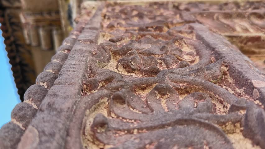 A downward tracking view reveals intricate ancient Indian stone carvings on a weathered fort wall, showcasing detailed patterns and centuries-old craftsmanship.