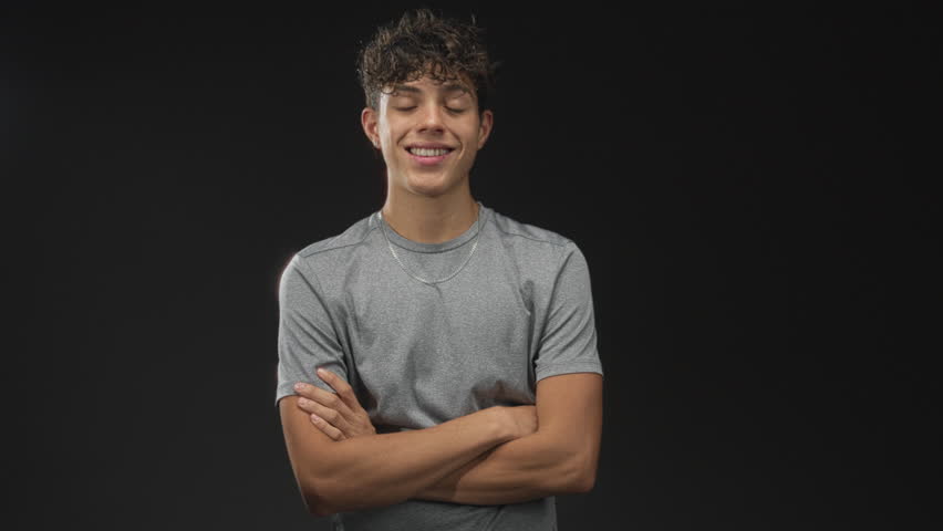 Boy wearing gray t shirt and silver necklace smiling with arms crossed in studio with black backdrop and soft lighting; youthful confidence.