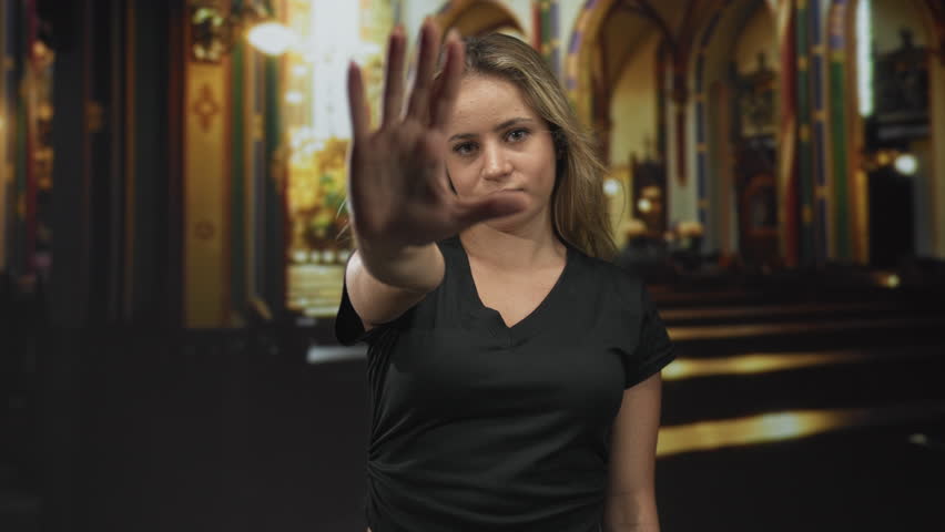 Blonde woman with outstretched hand showing palm forward and stern gaze in church building aisle; defiance conviction.
