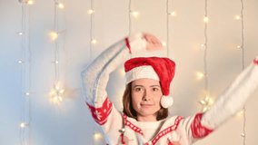 A young woman dancing on a festive white background with glowing holiday lights, playfully waving a Santa hat in her hand and enjoying the Christmas mood. - Powered by Shutterstock - Get 15% off with code: PIKWIZARD15