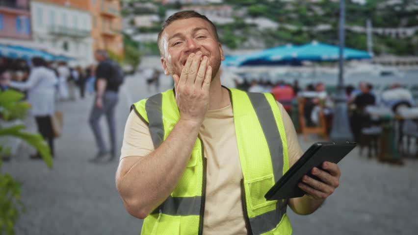 Man covering mouth with hand while laughing and holding tablet on street terrace; casual worksite amusement.
