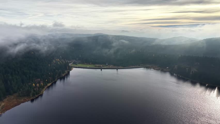 aerial misty reservoir with dam and bridge, still water reflecting pale sky, low clouds hugging forested shore