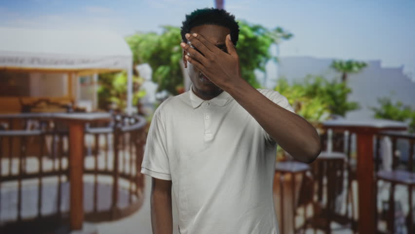 Man wearing white polo shirt with raised hand making stop gesture on building terrace by wooden railing under sunlight; caution boundary.