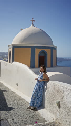 Woman leaning on a white wall beside a greek church building in santorini, shielding her eyes and looking up toward the sky; serenity travel.