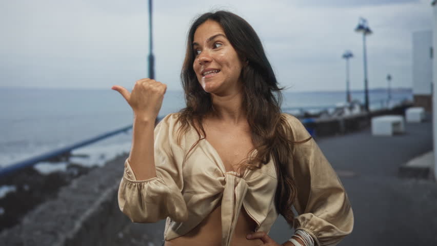 Woman showing cleavage and pointing thumb to the side on street promenade by the sea; joy holiday confidence.
