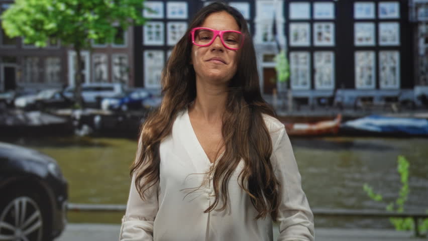 Woman wearing pink glasses forming a heart with hands on a street by an amsterdam canal, smiling; love connection.
