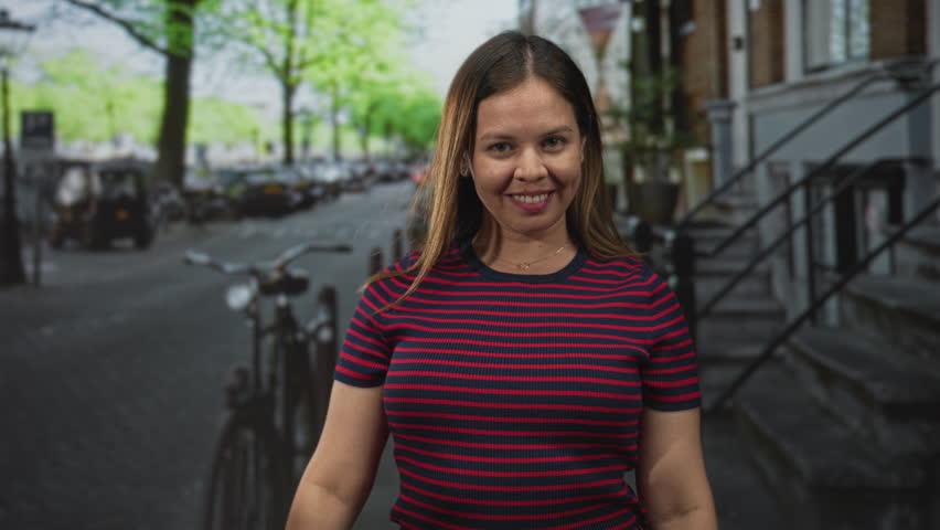 Woman points finger at camera on a city street with bicycles and stoops visible, striped shirt front clear; playful confidence.