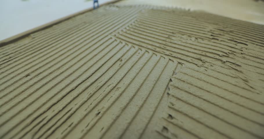Laying Ceramic Tiles. Troweling adhesive onto a concrete floor in preparation. Applying thin set tile mortar on a tile. A male tiler applies tile adhesive solution to the floor in a room. Close-up