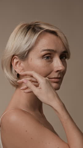 Vertical close up of blonde woman touching her delicate facial skin with manicured hand and smiling