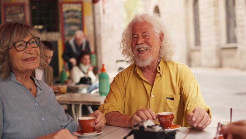 Cheerful elderly man and woman laughing and chatting together while drinking coffee at a sidewalk cafe table, enjoying their retirement