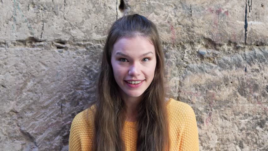 A cheerful girl with long flowing hair, wearing a bright yellow sweater, poses joyfully in front of a beautifully textured stone wall that reflects her vibrant and lively personality perfectly