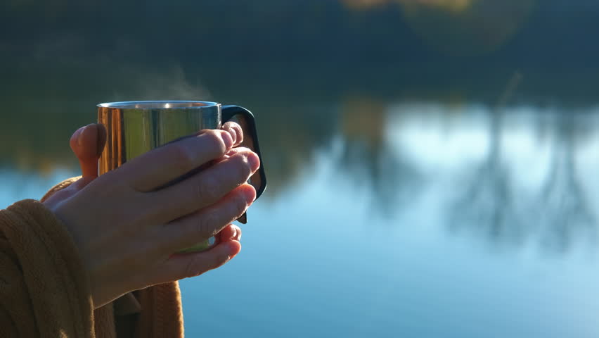 Woman holding steaming hot drink outdoors by lake. Female hands holding a metal mug with a hot steaming drink on a tranquil autumn morning, with the calm blue water of a lake and forest reflections