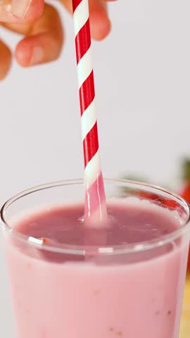 Hand stirs creamy pink fruit smoothie in glass using red striped straw, bright studio lighting