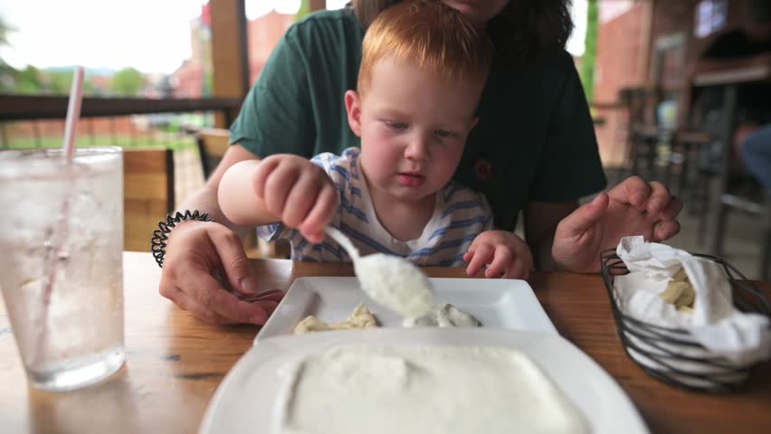 child learns selffeeding, toddler practices eating outdoors, young child develops selffeeding skills outside, child demonstrates emerging independence by selffeeding at outdoor dining area