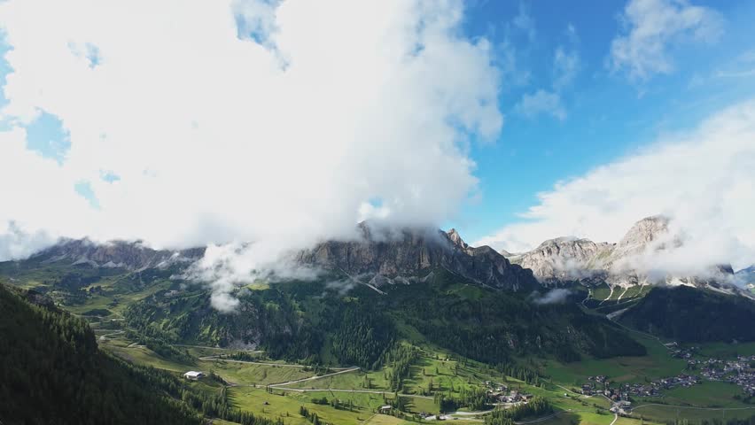 aerial dolomites panorama with clouds rolling over sunlit valley, green meadows and scattered chalets, dramatic peaks piercing mist, cinematic 4k sweep for travel and nature projects