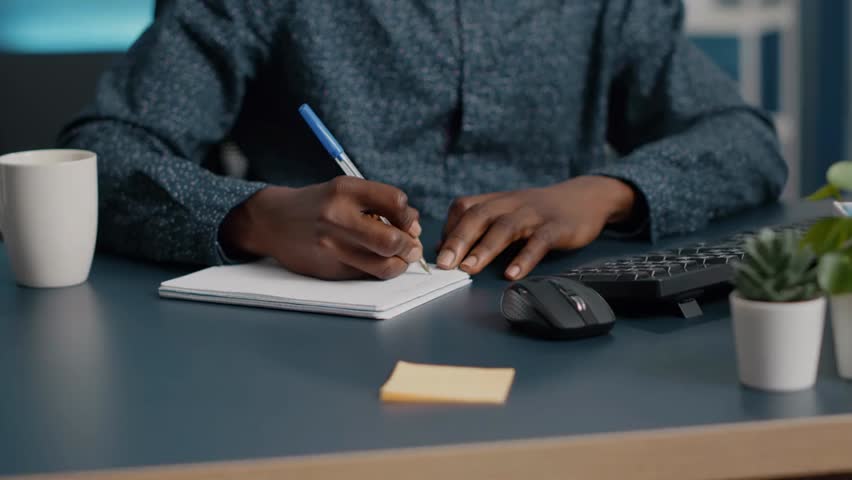 Close-up of African American man writing notes on a notepad, working or studying from home office, focusing on hands and pen.