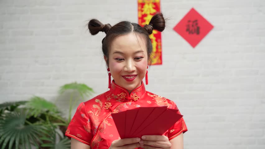 Happy young Asian woman in traditional red Cheongsam dress holding Ang Pao red envelopes for Chinese New Year celebration against white brick background