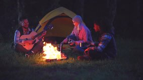 Cheerful multiethnic friends enjoying a summer night together, sitting on the grass around a campfire, singing songs while a man plays the acoustic guitar in front of a tent - Powered by Shutterstock - Get 15% off with code: PIKWIZARD15