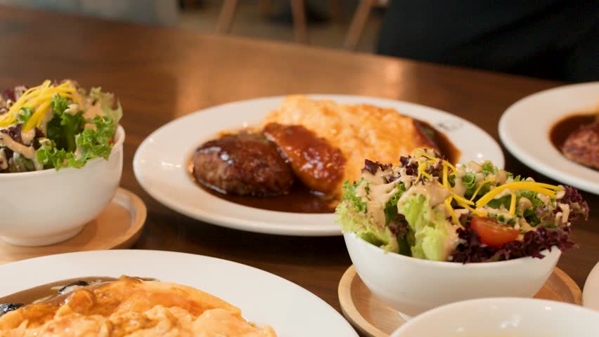 Hand places salad bowl beside omelette rice and hamburger steak under warm, natural restaurant lighting