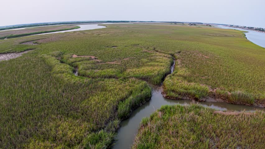 Drone footage captures green marshlands with winding waterways and tidal creeks near Charleston, South Carolina. The aerial view shows salt marsh vegetation and natural patterns under a pastel sky.