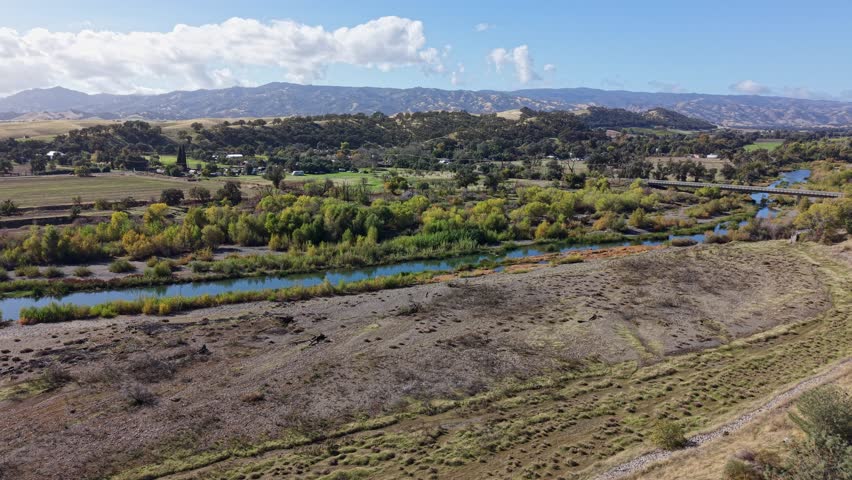 Drone slowly rises and drifts backward, revealing a calm river, green trees, a bridge, and distant rolling hills in Capay, California.