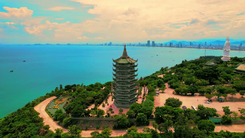 Cinematic aerial orbit shot around the Xa Loi Tower at Linh Ung Pagoda, Son Tra Peninsula, beautifully displaying the panoramic view of the vast blue sea and the distant modern cityscape of Da Nang, V