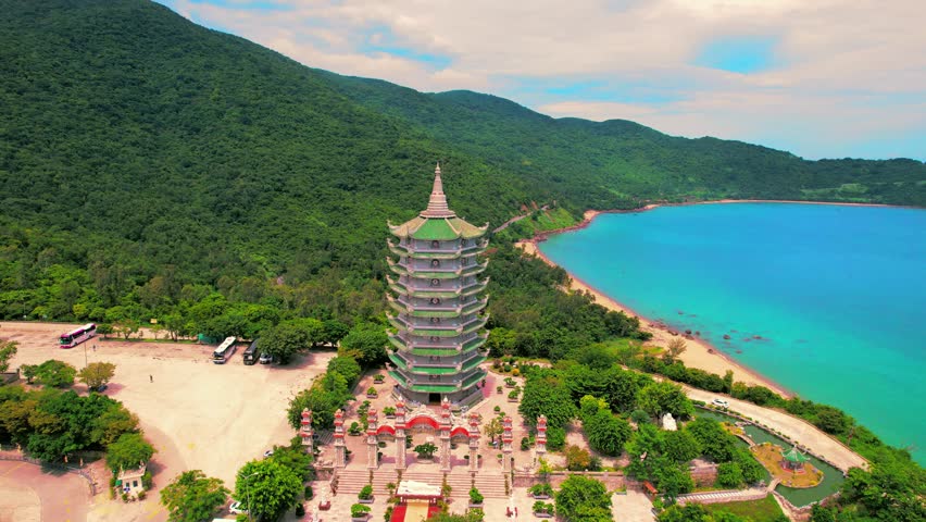 Cinematic aerial orbit shot showcasing the Xa Loi Tower at Linh Ung Pagoda, framed by the majestic Son Tra mountain range and the sweeping view of the Da Nang sea and coastline.