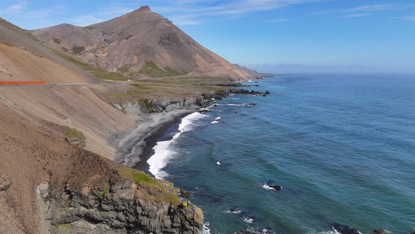 Aerial view of ocean waves hitting the rugged cliffside mountains of Iceland, revealing a winding coastal road, steep rocky slopes, and bright blue water stretching along the dramatic shoreline.