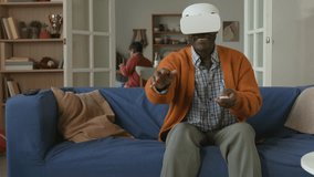 Smiling African American grandfather using VR headset to play video game sitting on couch in living room while young grandchildren interacting with devices in next room - Powered by Shutterstock - Get 15% off with code: PIKWIZARD15