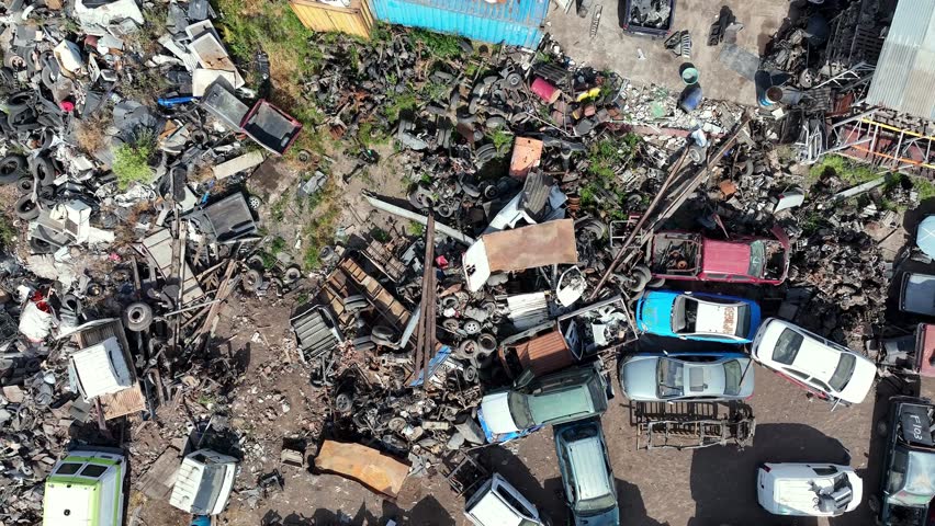 Forward drone aerial of chaotic junkyard with scrapped cars, trucks, auto parts, and metal debris piles in industrial salvage yard